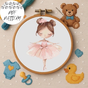May include: A cross-stitch pattern of a ballerina in a pink tutu, displayed in a wooden embroidery hoop. The image includes a teddy bear, a baby onesie, a pacifier, and a rubber duck. The text "PDF PATTERN" is visible.
