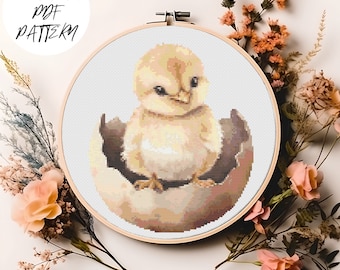 May include: A cross stitch pattern of a yellow chick hatching from a brown egg. The chick is sitting in the egg with its head tilted to the side and its eyes closed. The pattern is framed in a wooden hoop.