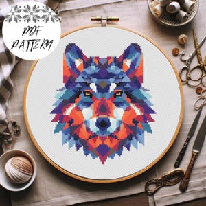May include: A colorful cross-stitch pattern of a wolf's head, featuring geometric shapes in shades of blue, orange, and purple. The design is set within a wooden embroidery hoop. The text "PDF PATTERN" is in a white circle in the upper left corner.