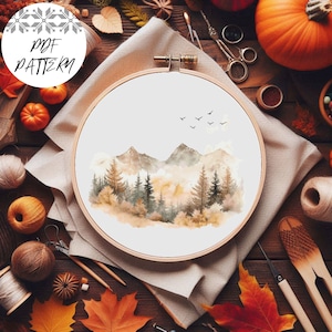 Puede incluir: Un patrón de punto de cruz de un paisaje de acuarela con montañas, árboles y pájaros volando en el cielo. El patrón se muestra en un aro de bordado de madera sobre una mesa de madera rústica con decoraciones de otoño.