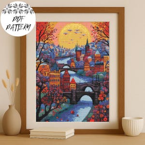 May include: A framed cross-stitch pattern of a cityscape with a river, bridges, and buildings in shades of red, orange, yellow, and blue. The text "PDF PATTERN" is in a white circle in the upper left corner. The artwork is in a wooden frame.