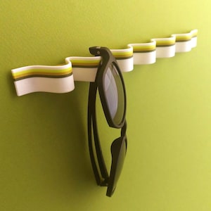 May include: A wall-mounted, wavy white storage strip featuring horizontal stripes in shades of olive green, yellow, and dark gray. The flexible, undulating design holds a pair of black-framed sunglasses hanging from one of its curved sections.
