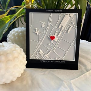 May include: A black framed map of a location in Sweden, with a red heart marking a specific point. The map is white and gray, with the text "Sweden - 2019/08" and coordinates. Two white, spherical candles are in the foreground.