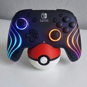May include: Black Nintendo Switch controller with colorful LED accents, resting on a red, white, and black Poke Ball-shaped stand. The controller has the Nintendo Switch logo.