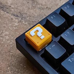 May include: A custom keyboard key, featuring a yellow question mark block design. The key is a replacement for a standard keyboard key and is set against a black keyboard. The question mark is white, and the key is a vibrant orange.
