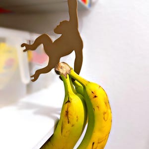 May include: A wooden monkey-shaped banana holder. The brown monkey silhouette hangs from a shelf, holding three ripe yellow bananas. The bananas have brown spots, indicating ripeness. The holder is designed to keep bananas fresh.