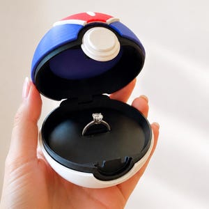 May include: A silver-toned ring with a single clear stone set in a prong setting, displayed inside a spherical, hinged container designed with a red, white, and blue color scheme, featuring a prominent white circular button on the front exterior.