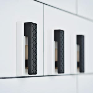 May include: Three vertical cabinet pulls mounted on white cabinet doors. Each pull features a rectangular black handle with a textured honeycomb pattern on the front surface, set against a solid white rectangular base plate for a clean contrast.