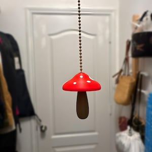 May include: A whimsical mushroom-shaped light fixture. The cap is bright red with white spots, and the stem is brown. It hangs from a brown beaded chain. The fixture is suspended in front of a white door.