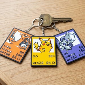 May include: Keychains featuring pixelated images of Charizard, Pikachu, and Mewtwo, resembling trading cards. The keychains are attached to a key. The Charizard keychain is orange, the Pikachu keychain is yellow, and the Mewtwo keychain is purple.