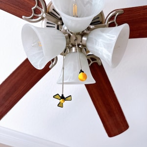 May include: A ceiling fan with four wooden blades and a light fixture. The blades are a dark brown color, and the light fixture has four white glass shades. The fan has a brushed nickel finish and a decorative pull chain.
