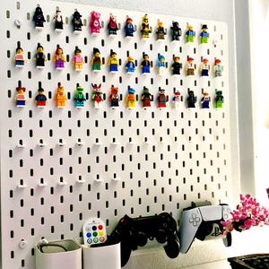May include: A white pegboard displays a collection of colorful Lego minifigures. Below, two white storage containers, a remote, and two game controllers are visible. The setup is on a white surface, with a touch of pink flowers.