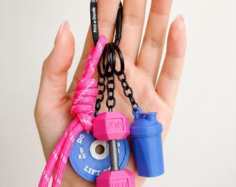 May include: A black carabiner keychain with the text Print-a-Doodle, featuring a bright pink paracord, a blue weight plate charm labeled 0 KG LIFT, a pink dumbbell charm labeled OLB, and a blue shaker bottle charm, all attached by black rings.
