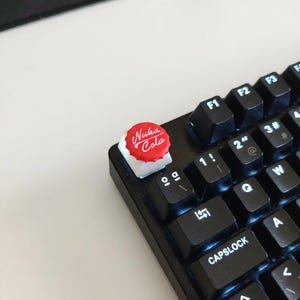 May include: A close-up of a black mechanical keyboard with a custom keycap. The keycap is red and white, resembling a bottle cap, with the words "Nuka Cola" printed on it. The keyboard keys are black.