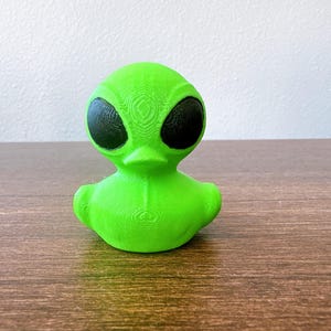 May include: A bright green alien-themed rubber duck with large black eyes. The duck has a textured surface and is sitting on a wooden table. The alien duck is a fun and unique novelty item.