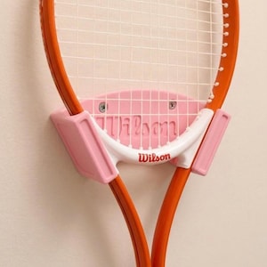 May include: An orange and white Wilson tennis racket mounted on a light pink plastic wall bracket. The racket features a white string pattern and the Wilson logo printed in red on the throat. The bracket holds the racket frame securely in place.