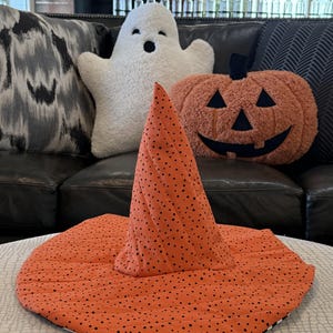 May include: A quilted orange witch hat featuring a black polka dot pattern. The hat has a tall, pointed crown and a wide, circular brim with visible stitching lines throughout the fabric, creating a textured appearance for this seasonal accessory.