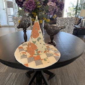 May include: A quilted witch hat featuring a cream-colored fabric with an orange rose floral pattern and a black and white houndstooth checkered border. The hat has a pointed crown and a wide brim, with small black spider motifs scattered throughout.