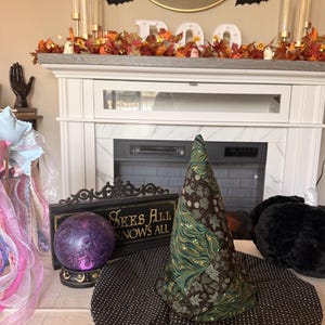 May include: A fabric witch hat featuring a conical top with a green and brown leaf pattern and a wide, flat brim with a black and white polka dot design. Next to the hat is a purple sphere on a stand and a black sign with the text SEES ALL KNOWS ALL.