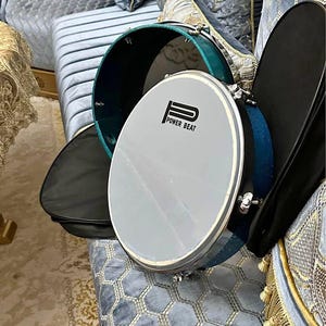Può includere: Un tamburo con una pelle grigia e il testo "POWER BEAT" è visualizzato. Il tamburo ha un bordo color verde acqua e hardware argentato. È visibile una custodia nera. Il tamburo è appoggiato su una superficie blu imbottita.