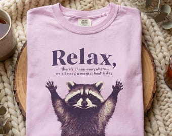 May include: A light purple t-shirt with the text "Relax, there's chaos everywhere... we all need a mental health day." A raccoon illustration is featured below the text with its arms raised. The shirt is on a wooden surface.