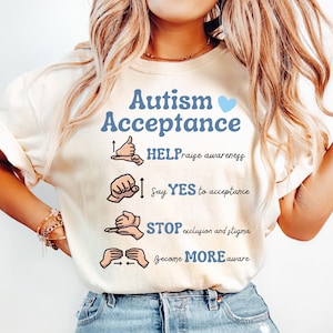 Puede incluir: Camiseta de color marfil con el texto "Autism Acceptance" y un corazón azul. La camiseta presenta una lista de símbolos de lenguaje de señas con el texto "HELP raise awareness", "Say YES to acceptance", "STOP exclusion and stigma", y "Become MORE aware".
