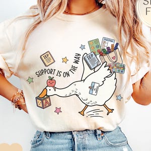May include: Ivory-colored t-shirt featuring a cartoon goose carrying school supplies. The goose has an apple on its beak and a block with the letter "A". Text on the shirt reads "Support is on the way".
