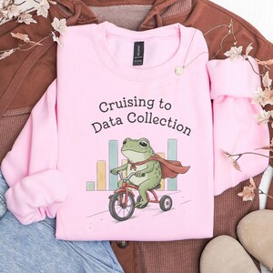 Könnte beinhalten: Ein hellrosa Sweatshirt mit Rundhalsausschnitt und einer Grafik eines grünen Frosches, der einen roten Umhang trägt und auf einem roten Dreirad fährt. Der Text "Cruising to Data Collection" steht darüber, ergänzt durch bunte Balkendiagramme.