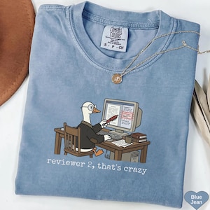 May include: A blue Comfort Colors t-shirt with a cartoon goose in a suit, working on a computer. The text "reviewer 2, that's crazy" is printed below the image. A gold necklace is draped on the shirt. The shirt is a light blue colour.