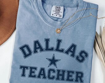 Op de afbeelding: Een blauw, kort mouw t-shirt met de woorden "DALLAS TEACHER" in marineblauwe blokletters en een ster. Een gouden ketting met een hanger ligt over het shirt. Het shirt is opgevouwen en heeft een label.
