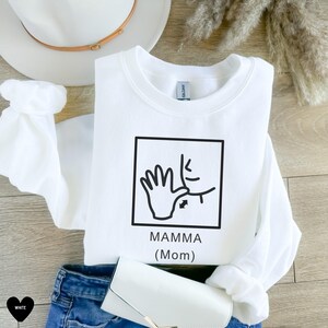 May include: White sweatshirt with a black graphic of a hand signing "mamma" in American Sign Language. The graphic is in a square with the text "MAMMA (Mom)" below it.
