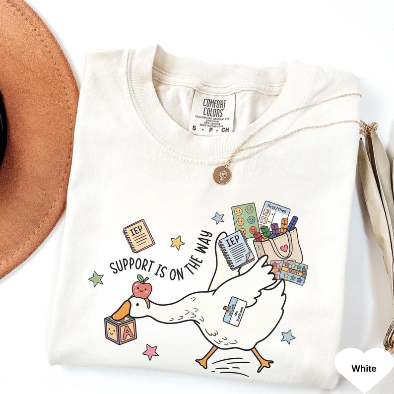 May include: Cream-colored t-shirt with a whimsical design featuring a cartoon goose carrying school supplies. The shirt has the text "Support is on the way." A gold necklace and a brown hat are also visible.