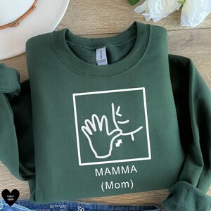May include: A dark green sweatshirt with a white graphic of a hand signing "mamma" in American Sign Language. The text "MAMMA (Mom)" is printed below the graphic.