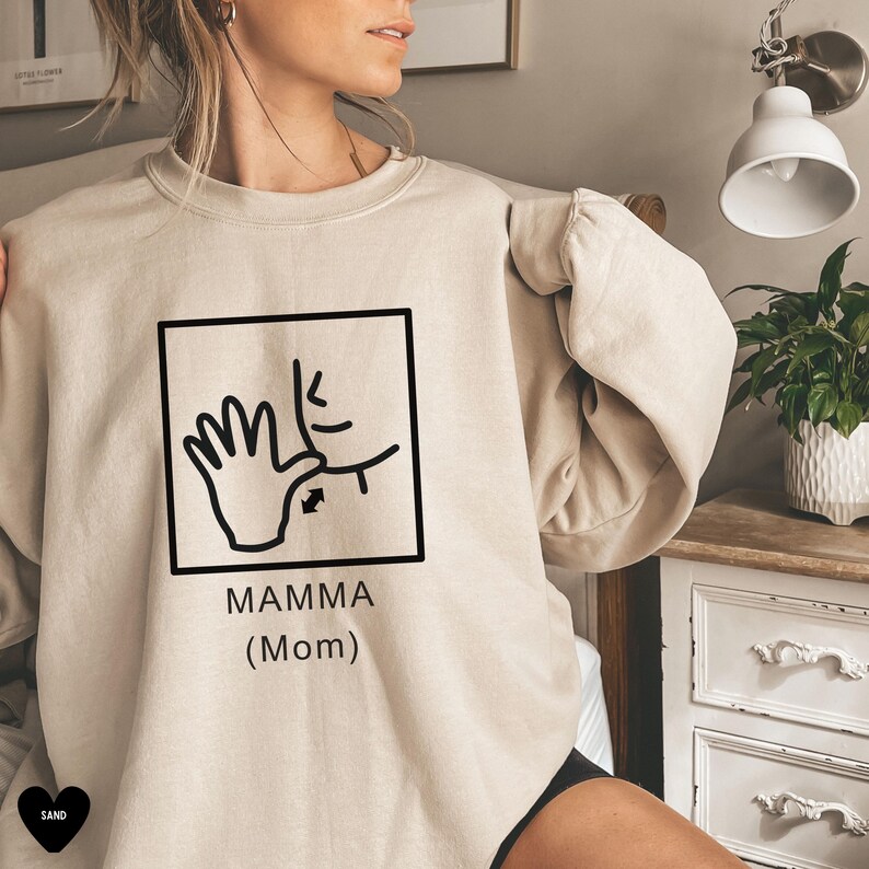 May include: A beige sweatshirt with a black graphic of a hand signing "mamma" in American Sign Language. The graphic is in a square with the text "MAMMA (Mom)" below it.