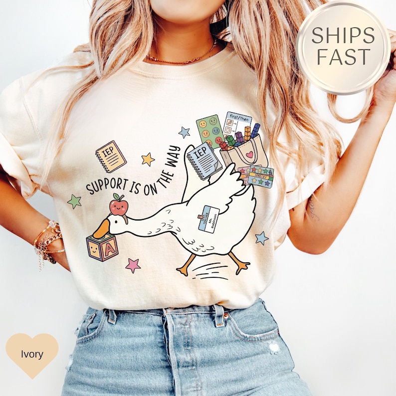May include: Ivory-colored t-shirt featuring a cartoon goose carrying school supplies. The goose has an apple on its beak and a block with the letter "A". Text on the shirt reads "Support is on the way".