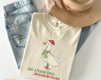 Op de afbeelding: Ivoor T-shirt met een feestelijke origami gans met een kerstmuts en sjaal. Op het shirt staat de tekst "Just a Festive Goose... Unfolding My Feelings". Het shirt wordt gecombineerd met een lichtblauwe jeans en een beige hoed.