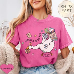 May include: A pink t-shirt with a graphic of a white goose carrying a bag of school supplies. The goose has a name tag and is walking towards the right. The text "SUPPORT IS ON THE WAY" is printed on the shirt.