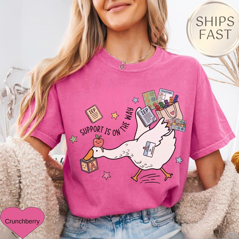 May include: A pink t-shirt with a graphic of a white goose carrying a bag of school supplies. The goose has a name tag and is walking towards the right. The text "SUPPORT IS ON THE WAY" is printed on the shirt.