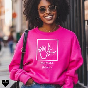 May include: Pink sweatshirt with a white graphic of the American Sign Language sign for "mom" and the text "MAMMA (Mom)".