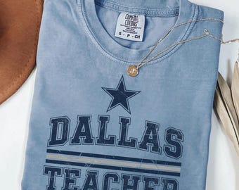 Op de afbeelding: Een blauw spijkerbroekkleurig t-shirt met de woorden "DALLAS TEACHER" in een verweerde letter, met een marineblauwe ster boven het woord "DALLAS". Een gouden ketting met een hanger is over het shirt gedrapeerd.