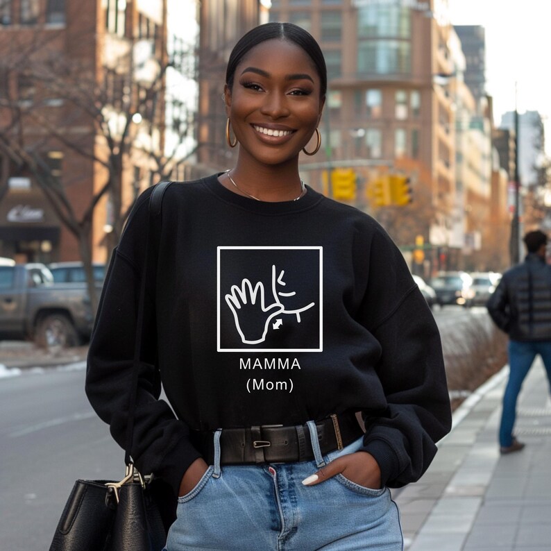 May include: A black sweatshirt with a white graphic of a hand signing the word 'mamma' in American Sign Language. The text 'MAMMA (Mom)' is printed below the graphic.