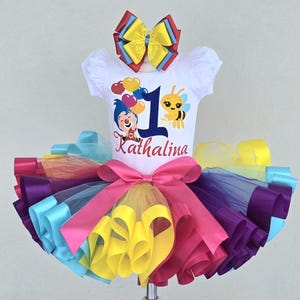 May include: A white short-sleeve shirt featuring a cartoon character, a bee, heart balloons, the number 1, and the name Kathalina, paired with a multi-colored tulle tutu skirt with a large pink bow and a matching multi-colored hair bow accessory.