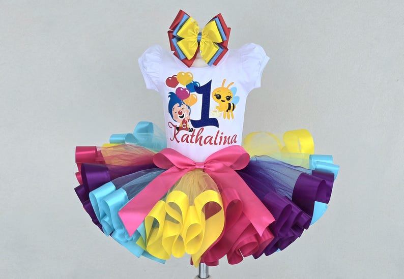 May include: A white short-sleeve shirt featuring a cartoon character, a bee, heart balloons, the number 1, and the name Kathalina, paired with a multi-colored tulle tutu skirt with a large pink bow and a matching multi-colored hair bow accessory.