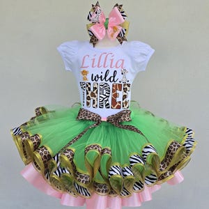 May include: A white short-sleeved shirt with the text Lillia wild THREE and animal graphics, paired with a layered green tulle tutu skirt featuring leopard and zebra print ribbon trim, and a matching pink and animal print hair bow accessory.
