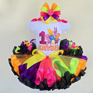 May include: A colorful tutu set for a child's first birthday. The white t-shirt features a number 1 and cartoon characters. The tutu has layers of pink, orange, green, purple, and black ribbons. A large bow completes the outfit.