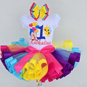 May include: A white short-sleeved shirt featuring a blue number one, a cartoon clown, a bee, and the name Kathalina, paired with a multi-colored tulle tutu skirt in layers of pink, purple, yellow, and blue, topped with a matching hair bow.