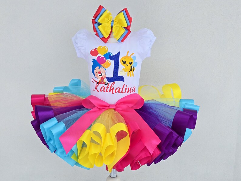 May include: A white short-sleeved shirt featuring a blue number one, a cartoon clown, a bee, and the name Kathalina, paired with a multi-colored tulle tutu skirt in layers of pink, purple, yellow, and blue, topped with a matching hair bow.