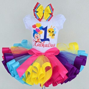 May include: A white short-sleeved shirt featuring a graphic of a cartoon character, a bee, and balloons with the number 1 and the name Kathalina. The shirt is paired with a multi-colored tulle tutu skirt in pink, yellow, purple, and blue with a large bow.