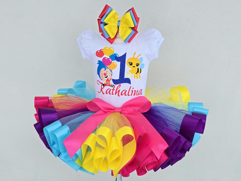May include: A white short-sleeved shirt featuring a graphic of a cartoon character, a bee, and balloons with the number 1 and the name Kathalina. The shirt is paired with a multi-colored tulle tutu skirt in pink, yellow, purple, and blue with a large bow.