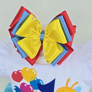 May include: A large multi-layered hair bow featuring vibrant red, blue, and yellow satin ribbons. The center is secured with a blue ribbon band, and the top yellow layer is embellished with small clear rhinestones along the side edges.